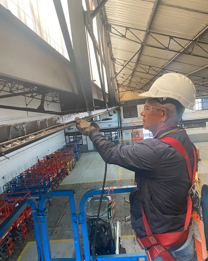 Técnico da JW com capacete, óculos e cinto de segurança limpando calha de galpão em plataforma elevatória