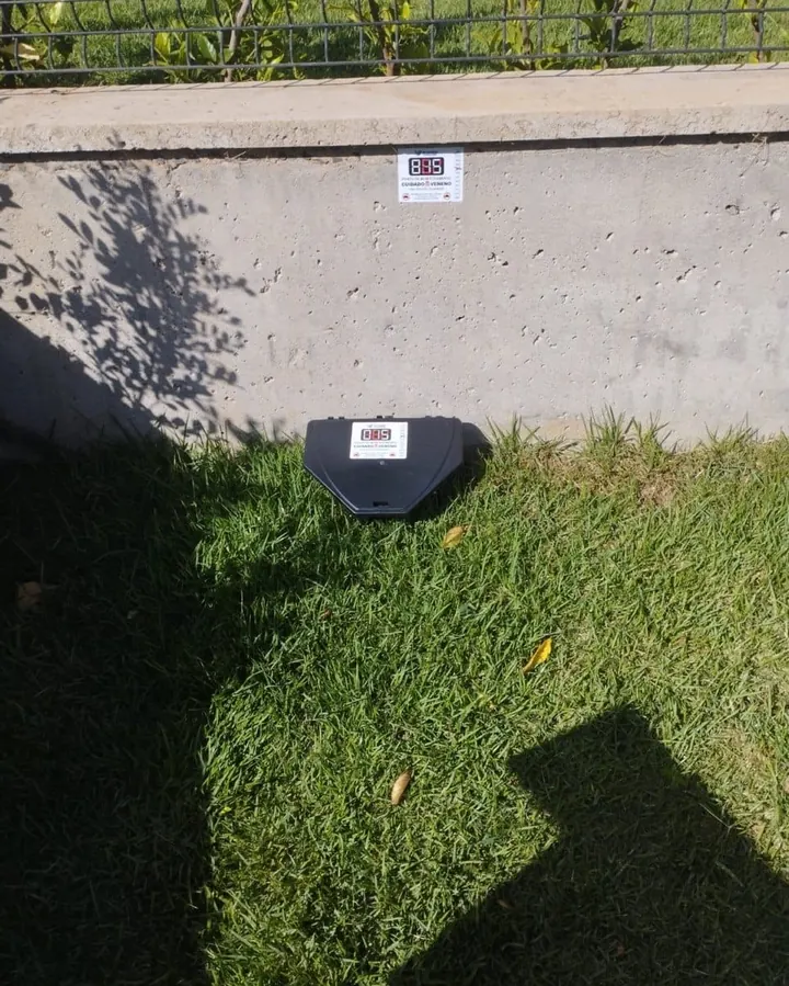 Porta-isca identificado instalado pela JW na base de um muro, com gramado limpo
